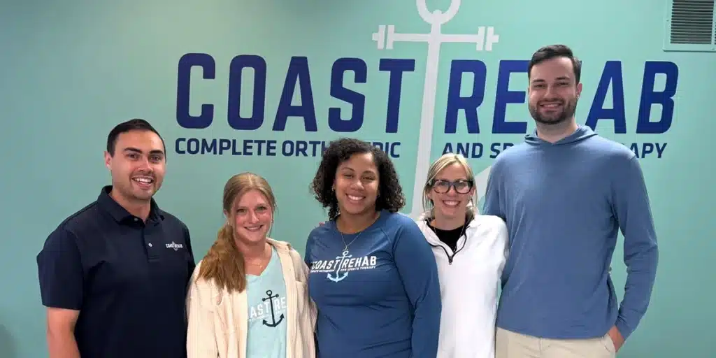 Five smiling Coast Rehab team members standing in front of the Coast Rehab Complete Orthopedic and Sports Therapy sign